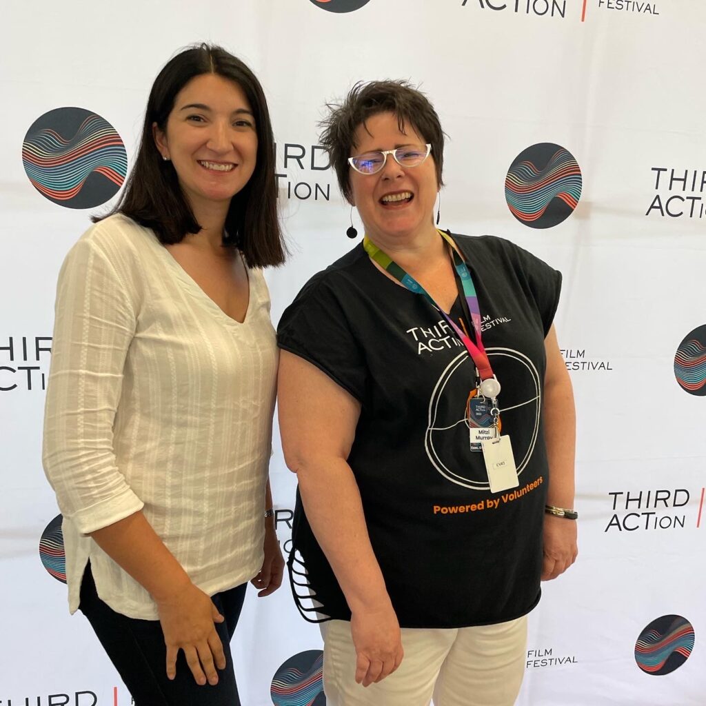 Two women posing in front of a sign for the Third Action Film Festival.