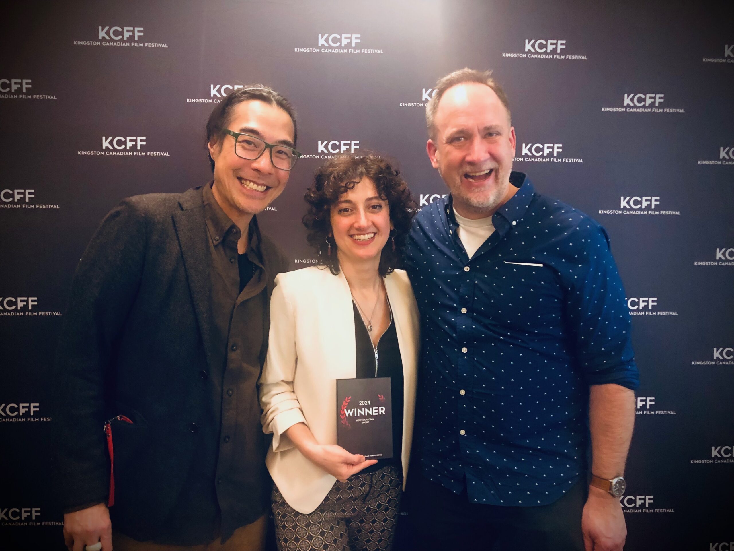 three people posing in front of a logo wall. The woman in the middle of the two men is holding an award that says Best Canadian Short